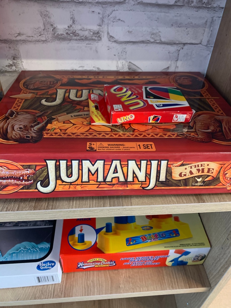 A shelf of board games, in the centre of the frame is Jumanji,  on top of that is  Uno  